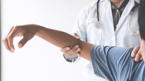 Eine Person mit Arztkittel und umgehängtem Stethoskop untersucht den Arm eines Patienten, indem sie Schulter und Ellbeuge fasst und augenscheinlich leicht bewegt.