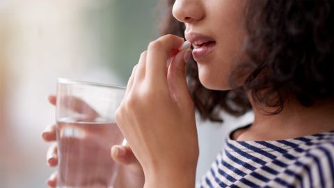 Acil kontrasepsiyon: genç bir kadın bir elinde bir bardak su tutuyor. Diğer eliyle yarı açık ağzına bir tablet tutuyor.