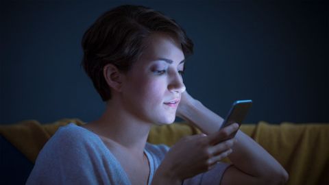 Young woman sitting on the sofa in semi-darkness. She is holding a mobile phone close to her face and looking at the screen with concentration. Her face is illuminated by the bluish light of the display.