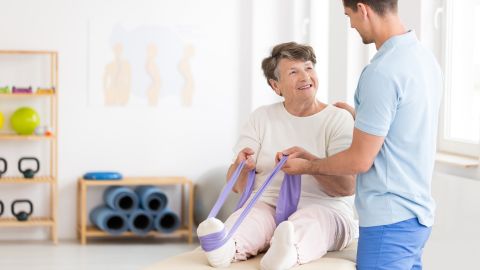 Osteoporosis: older woman sitting on a mat with her legs stretched out. The older woman has a fitness band stretched around one foot which she is pulling with both hands. A trainer is standing next to the woman and supporting her at her back and one hand.
