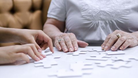 Home care for family members with dementia: the hands of an older woman and a younger person touching large jigsaw pieces spread out on a tabletop.