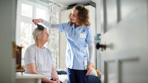 An older woman is sitting on the edge of a bed. A young woman is standing next to her. She is combing the hair of the older woman, who is clearly a care recipient.