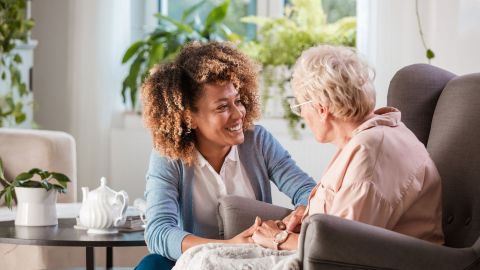 In need of care: older woman sitting in a chair. A younger woman is kneeling down next to her. She is holding the older woman’s hand in both hands, looking at her and smiling. Indoor plants in the background.