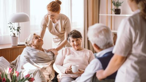 Vollstationäre Pflege im Heim: Zwei ältere Frauen und ein älterer Mann sitzen beisammen in einem Seniorenheim und unterhalten sich. Zwei Pflegerinnen stehen bei den Senioren.
