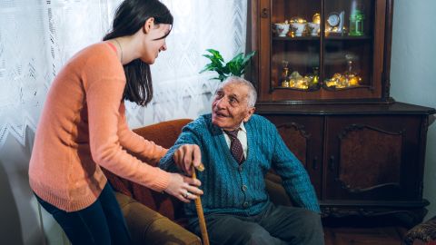 Older gentleman using his walking stick to try to get up out of his armchair. A young woman is helping him to stand up and supporting him by holding his arm.