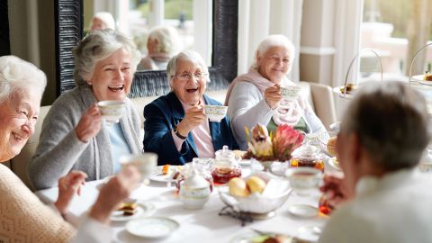Care group homes, where people live together and receive outpatient care: five older people sitting at a dining table with bowls of bread and jam. All the women have a cup in their hand which they are lifting up to toast each other.