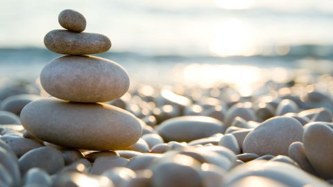 Vier Kieselsteine in unterschiedlicher Größen, die aufeinander gestapelt sind, wahrscheinlich an einem Strand. Im Hintergrund eine untergehende Sonne, deren Strahlen in Wasser reflektiert werden.