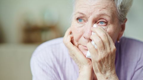 Psychische Gesundheit im Alter: Eine ältere Frau guckt nachdenklich geradeaus. Ihren Kopf hat sie auf eine Hand gestützt, mit der anderen Hand hält sie ein Taschentuch an ihr Gesicht. Sie wirkt traurig.