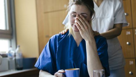 Mental health in the workplace: nurse sitting at a table with a cup. She is supporting herself on her left elbow and rubbing her hand across her face. A colleague is placing her hand on her shoulder and comforting her.
