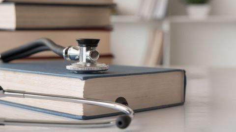 Ein Lehrbuch für Medizinrecht und ein Stethoskop liegen auf einem Tisch. Im Hintergrund weitere Bücher, die gestapelt auf dem Tisch liegen.