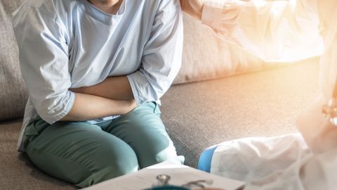 Irritable bowel syndrome: woman sitting on a couch holding her arms together and pressing them to her belly. The woman is leaning her chest forward slightly and pressing her thighs together. Her tension is obvious from the way she is holding her body.