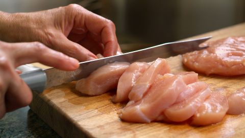 Die Hände einer Frau schneiden Hühnchenfleisch in Scheiben. Das Fleisch liegt auf einem hölzernen Schneidebrett.