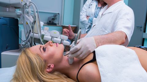 Hyperthyroidism: young woman lying on a treatment table at a doctor’s practice. A doctor wearing a surgical glove is holding a scanner to the woman’s larynx. The doctor is conducting an ultrasound scan of her thyroid gland.
