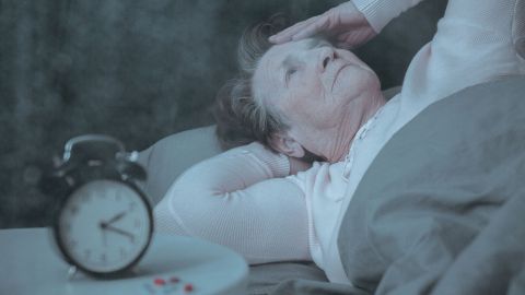 Sleep disturbances: older woman lying in bed clutching her head and looking at the ceiling. There is an alarm clock next to her on the bedside table.