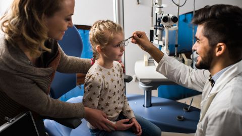 Amblyopie: Ein Mädchen sitzt auf einem Behandlungsstuhl in einer Praxis. Vor ihr kniet ein Augenarzt. Mit einer Hand hält er eine Brille vor die Augen des Mädchens. Eine Frau steht daneben und stützt den Oberkörper des Kindes.