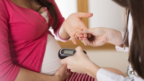 Pregnant woman sitting in the treatment room of a medical practice. A doctor is sitting opposite her. She is reaching for the pregnant woman’s hand with one hand. She is holding a glucose tolerance testing device in the other hand.