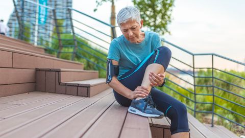 Tendinitis: older woman in sports clothing sitting on steps in the open air. She is holding her ankle and grimacing with pain.