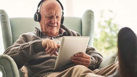 Older man sitting on a chair with outstretched legs. He is wearing headphones and smiling as he looks at the image on a laptop he is holding in his left hand. The man is touching the screen with the forefinger of his other hand.