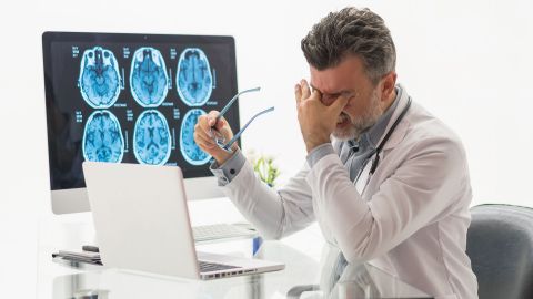 Treatment errors: doctor sitting in front of a laptop. There is a large screen next to him showing images of a scanned cranium. The doctor is pressing two fingers to his eyes, grimacing and showing his teeth which are tightly shut.