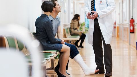 Ein Mann mit Gips am Bein und eine Frau sitzen in einem Wartebereich. Neben ihnen steht ein Arzt. Sie sprechen miteinander.