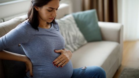 Типичные симптомы беременности. Молодая женщина на большом сроке беременности сидит на диване с закрытыми глазами. Она держит одну руку на животе, а другую — на спине. Похоже, что у нее болит спина.