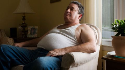 Overweight man sitting in a chair. A fat belly can be seen under the ribbed undershirt the man is wearing.