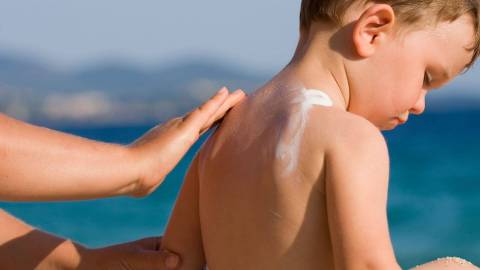 Маленький ребенок сидит на пляже, и ему на спину наносят солнцезащитный крем.