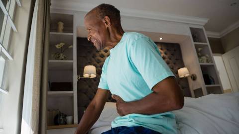 A middle-aged man is sitting on the edge of a bed and holding the right side of his abdomen. His facial expression indicates acute pain.