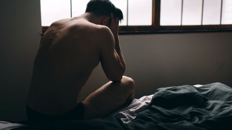 Premature ejaculation: young man sitting on the edge of a bed. His chest is bent forward, his elbows are placed on his thighs and he is holding his head in both hands.