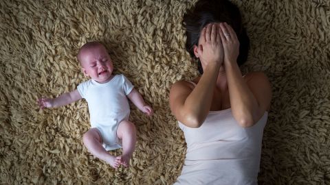 Eine Frau liegt auf einem Teppich und hält sich beide Hände vors Gesicht. Sie wirkt erschöpft. Neben ihr liegt ein Baby. Es scheint zu strampeln und zu weinen.