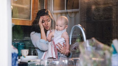 Wochenbettdepression: Eine gestresst wirkende Frau steht in der Küche, hält ein Baby im Arm und hält sich den Kopf.
