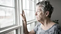 Eine ältere Frau lehnt am Fenster und schaut betrübt nach draußen.