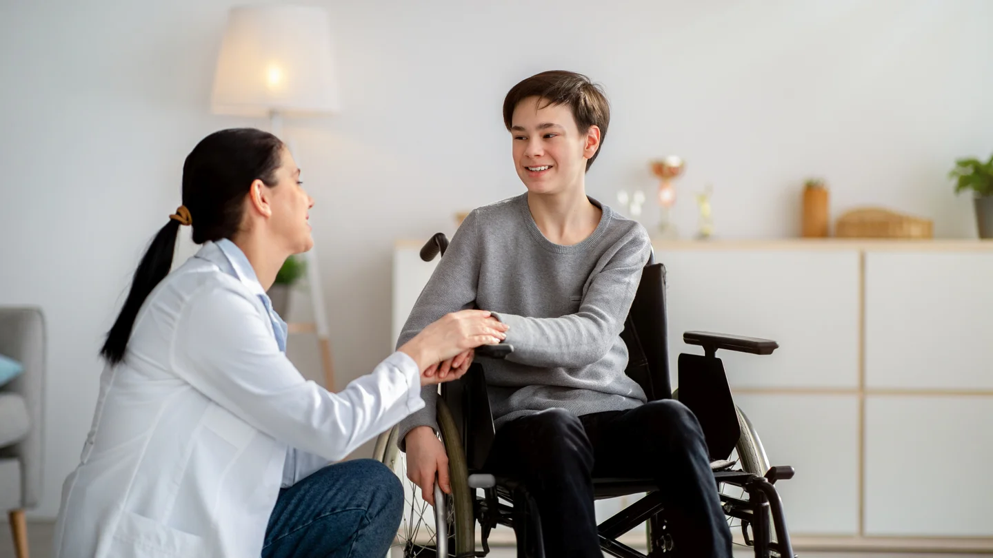 Ein Ärztin kniet neben einem Jungen im Rollstuhl und reicht ihm die Hand.