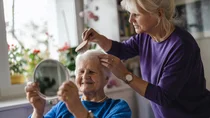 Eine Frau kämmt ihrer älteren Mutter die Haare. Die Mutter betrachtet sich dabei lächelnd im Spiegel.