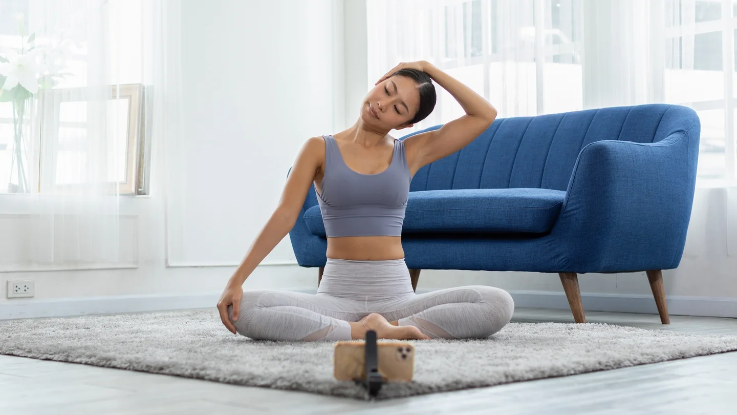 Eine Frau sitzt auf dem Boden vor ihrem Smartphone und macht Entspannungs- und Dehnungs&uuml;bungen. 