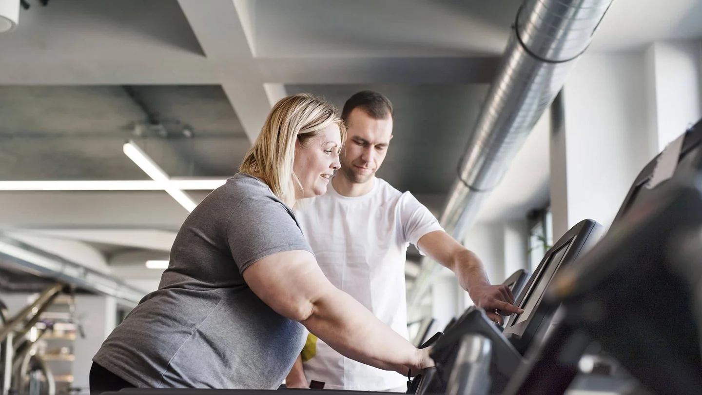 Ein Mitarbeiter eines Fitnessstudios zeigt einer Frau mit starkem Übergewicht, wie das Laufband funktioniert.