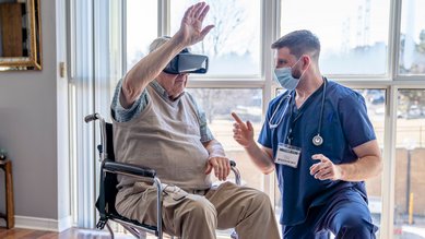 Ein im Rollstuhl sitzender Senior tr&auml;gt eine Virtual-Reality-Brille und hebt einen Arm. Er wird dabei von einem Therapeuten angeleitet, der neben dem Rollstuhl kniet.