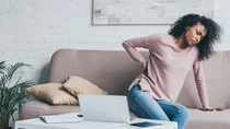 Eine Frau sitzt auf einer Couch. Mit einem Arm stützt sich die Frau ab, mit der anderen Hand fasst sie sich an den Rücken.