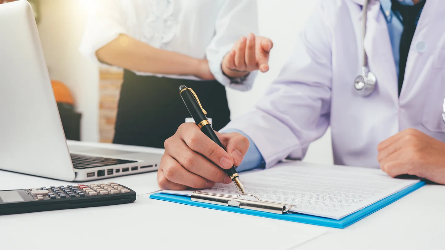 IGeL-Leistungen: Ein Arzt sitzt am Schreibtisch und füllt ein Formular aus. Eine Frau mit weißer Bluse steht hinter ihm. Sie hat den rechten Arm vor dem Bauch verschränkt und mit dem anderen Arm macht sie eine Handbewegung in Richtung des Arztes.