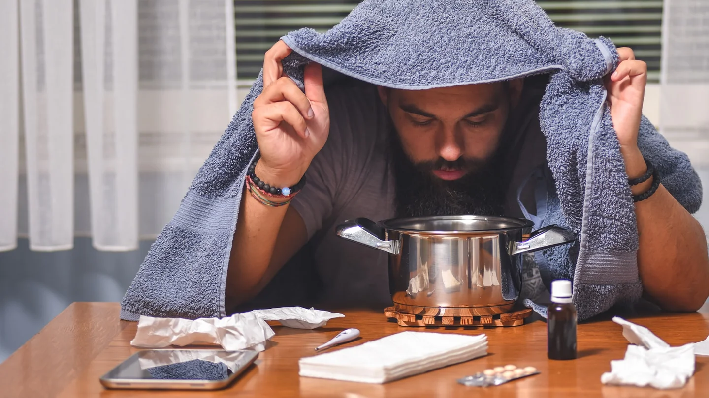 Ein Mann sitzt an einem Tisch, auf dem Taschentücher, Fieberthermometer, Tabletten, Nasentropfen und ein Topf stehen. Der Mann hält ein Handtuch über seinen Kopf und beugt sich über den Topf.