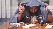Ein Mann sitzt an einem Tisch, auf dem Taschentücher, Fieberthermometer, Tabletten, Nasentropfen und ein Topf stehen. Der Mann hält ein Handtuch über seinen Kopf und beugt sich über den Topf.