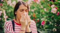 Eine Frau im T-Shirt schnäuzt sich in ihr Taschentuch. Im Hintergrund sind blühende Blumen zu sehen.