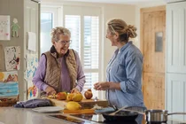 Eine Frau bereitet in der K&uuml;che eine Mahlzeit zu und unterh&auml;lt sich dabei mit einer Seniorin.