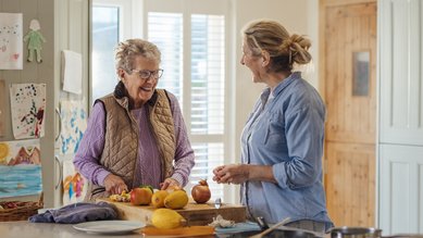 Eine Frau bereitet in der K&uuml;che eine Mahlzeit zu und unterh&auml;lt sich dabei mit einer Seniorin.