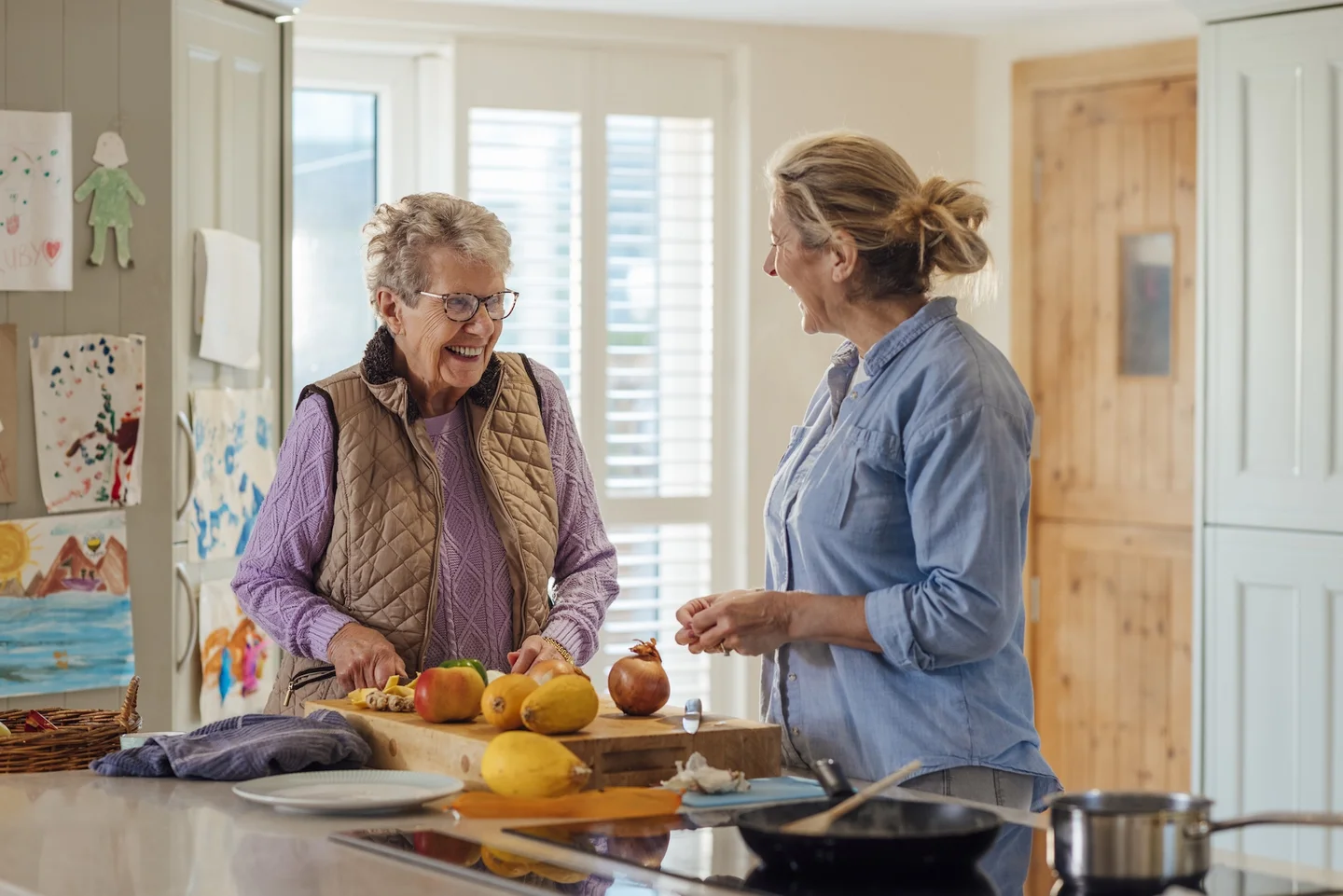Eine Frau bereitet in der K&uuml;che eine Mahlzeit zu und unterh&auml;lt sich dabei mit einer Seniorin.