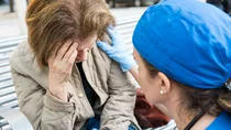 Eine Frau sitzt auf einer Bank, fasst sich an die Stirn. Eine Arzthelferin kniet vor ihr und fasst ihr mit der rechten Hand an die linke Schulter.