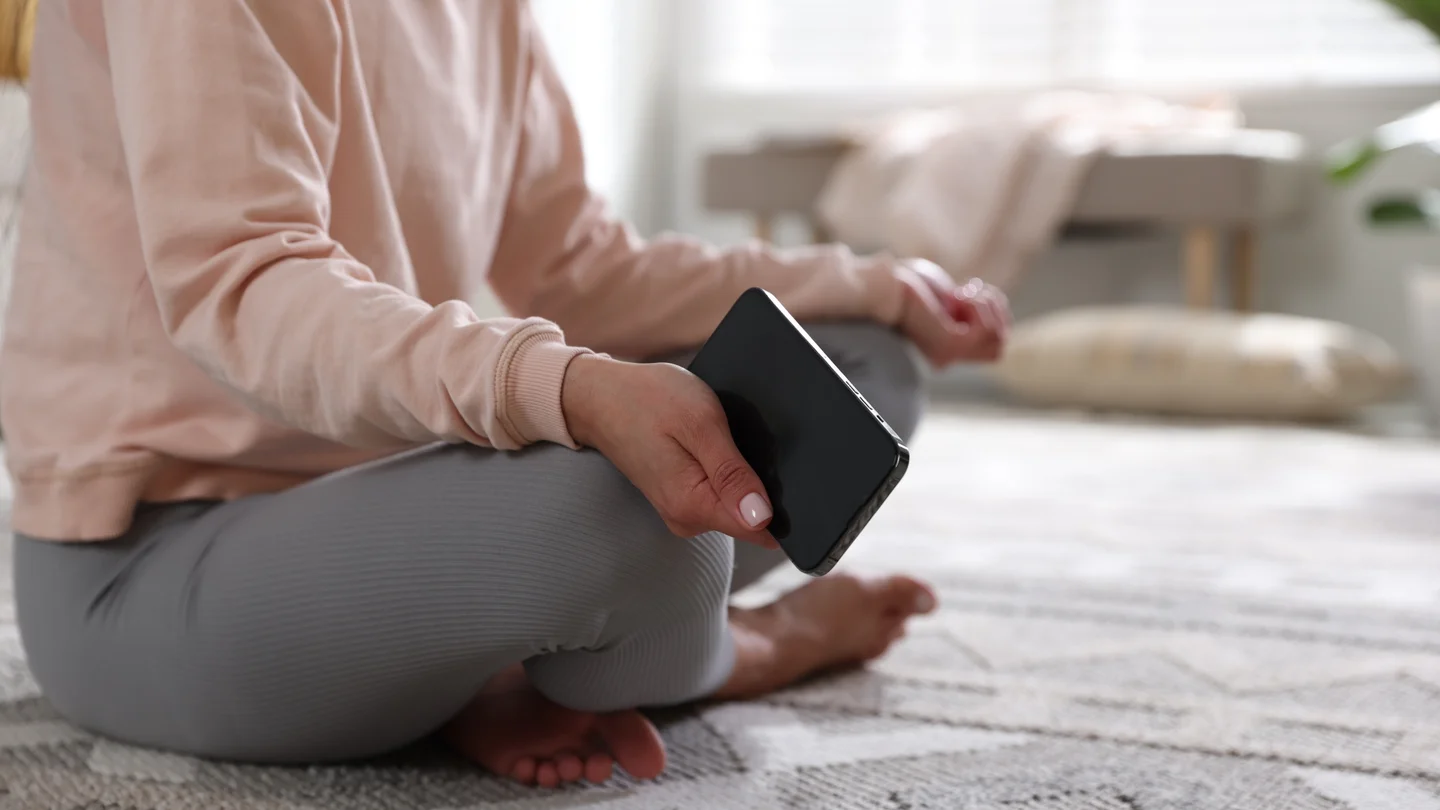 Eine Frau sitzt im Schneidersitz auf dem Boden und h&auml;lt ein Smartphone in der rechten Hand. 