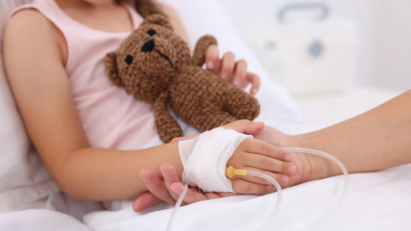 Ein M&auml;dchen liegt in einem Krankenhausbett und h&auml;lt einen Teddy in der Hand. Eine erwachsene Person h&auml;lt die Hand des M&auml;dchens, in der sich ein medizinischer Zugang befindet.
