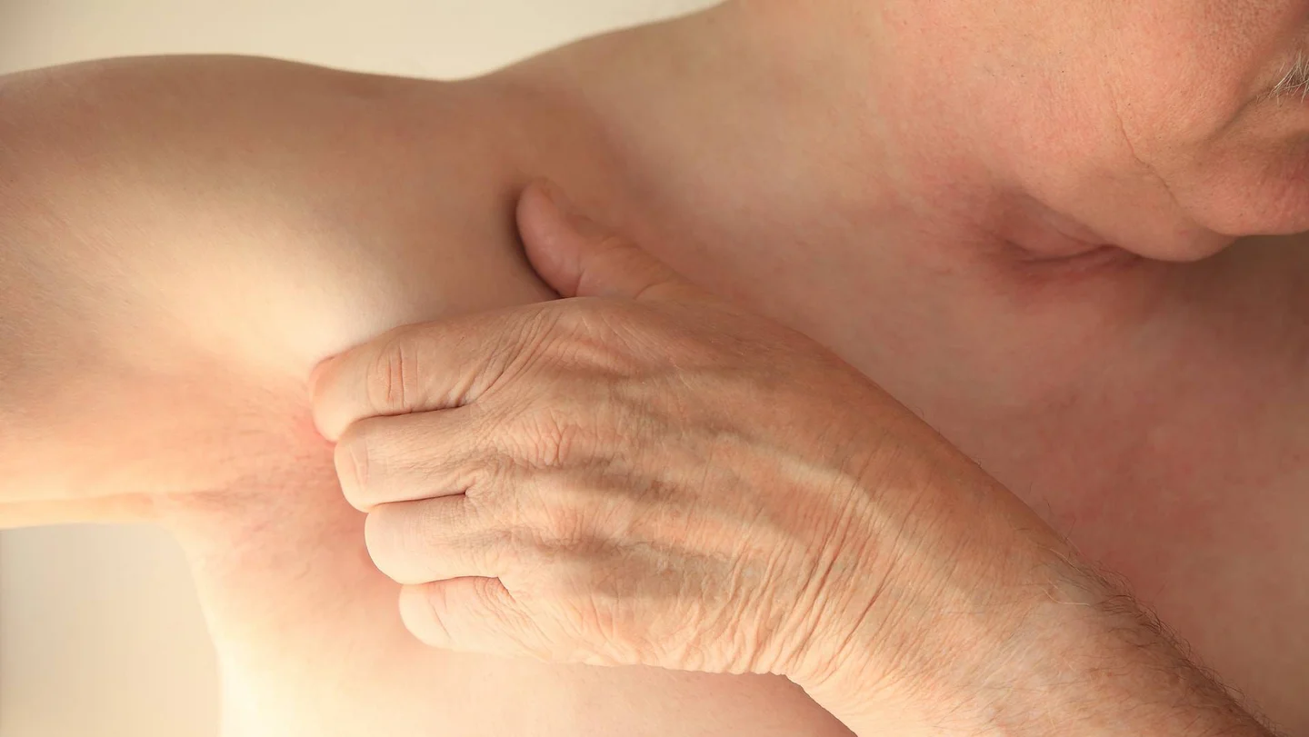 Ein Mann mit freiem Oberkörper streckt seinen rechten Arm zur rechten Seite aus. Mit der linken Hand umgreift er seine Achselhöhle.