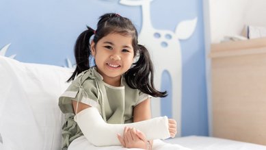 Ein M&auml;dchen sitzt mit einem eingegipsten Arm im Bett und l&auml;chelt.